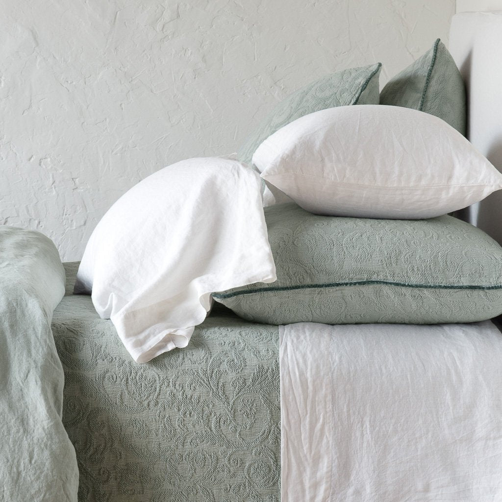 : Adele shams in eucalyptus on a bed shown from the side, layered with white sheets and pillowcases and a eucalyptus coverlet against a plain white wall.
