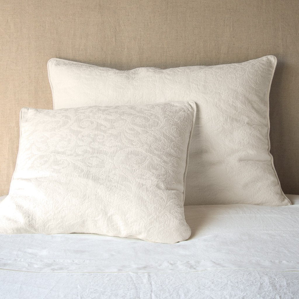 Parchment: Two shams , shown in euro and deluxe sizes, leaning against a neutral toned headboard on white bedding.