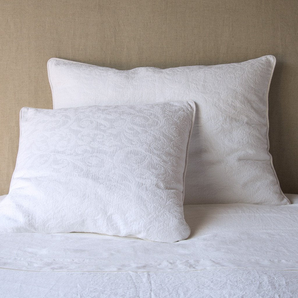 White: Two shams , shown in euro and deluxe sizes, leaning against a neutral toned headboard on white bedding.