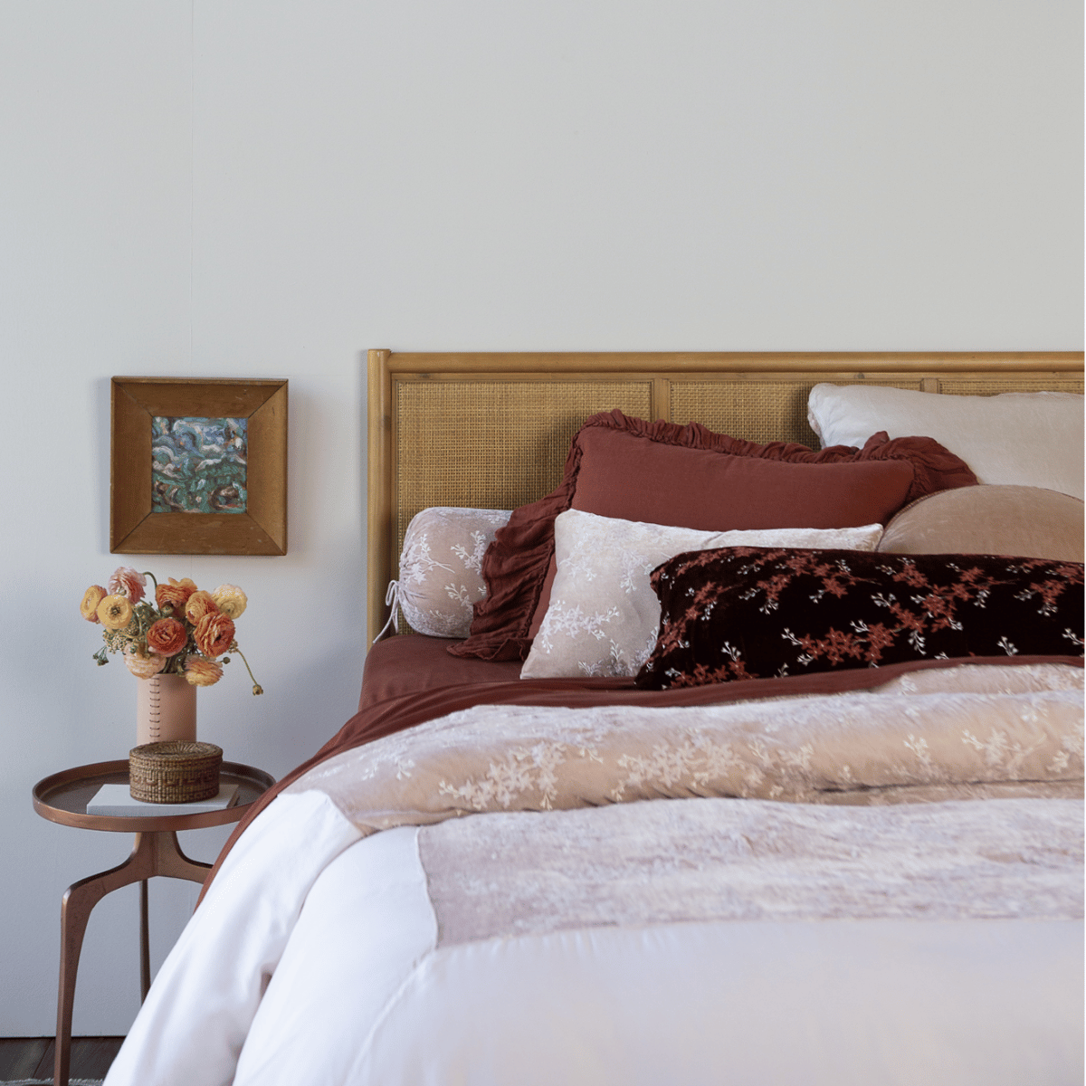 a pale pink duvet cover with dark terrcotta colored sheets and pillows against a rattan headboard with coordinating side table - shot from the foot of the bed.