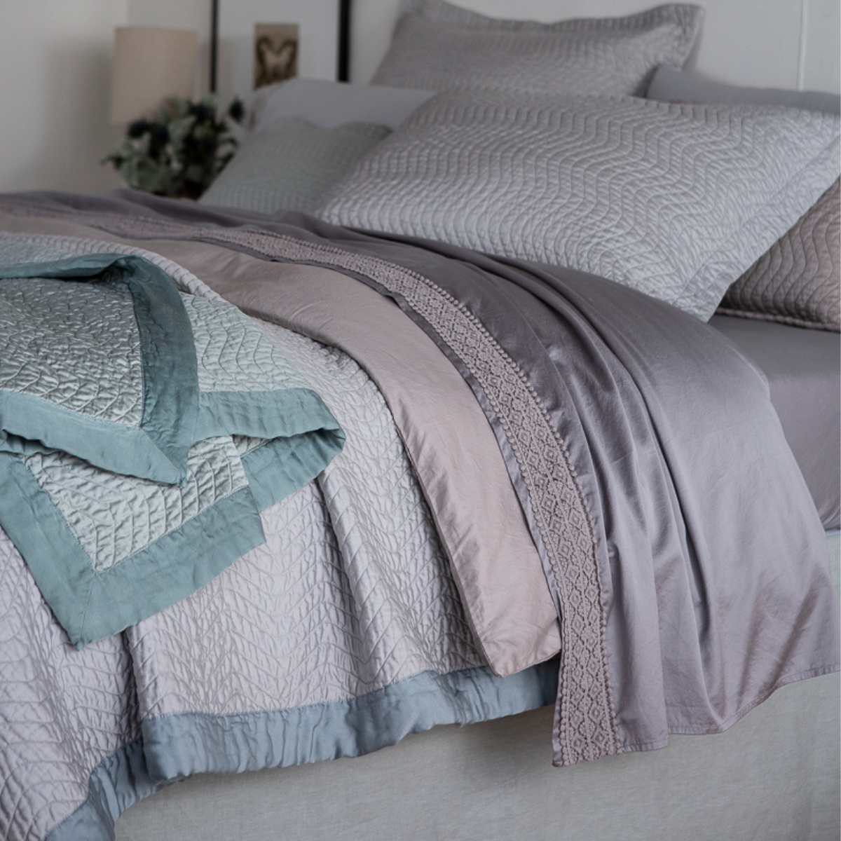 : a close up of the side edge of a bed dressed in cotton sateen. The shams, coverlet and throw blanket are quilted and the unquilted duvet cover and flat sheets are also visible. There is a band of cotton lace on the flat sheet's cuff.