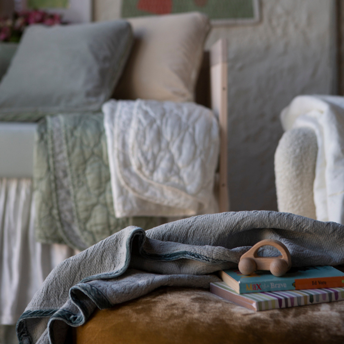 : Blanket in mineral draped over pouf, shown with books and toys in a green and cream toned nursery scene.