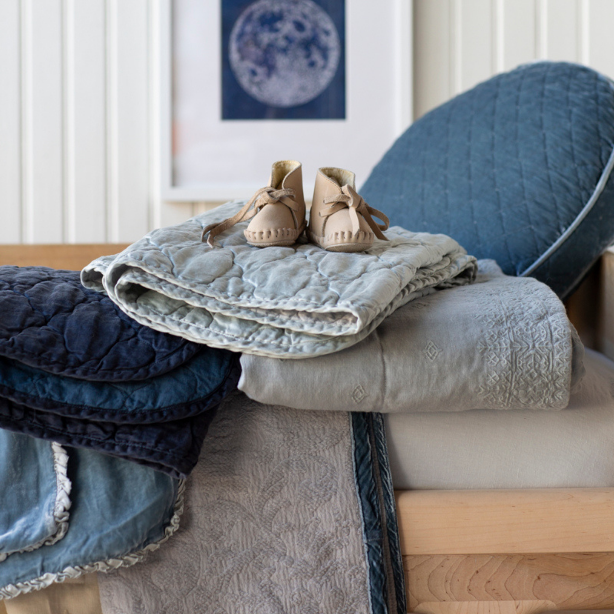 : Blanket in cloud, neatly folded, arranged with other blankets in a range of textures and blue tones on a light wood tone toddler bed.