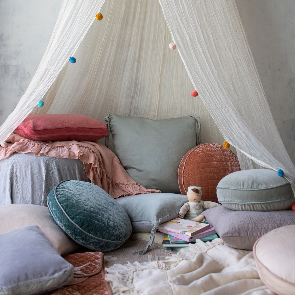 : Blanket in rouge folded and layered with many blankets and pillows in a cozy, canopy-covered reading nook.
