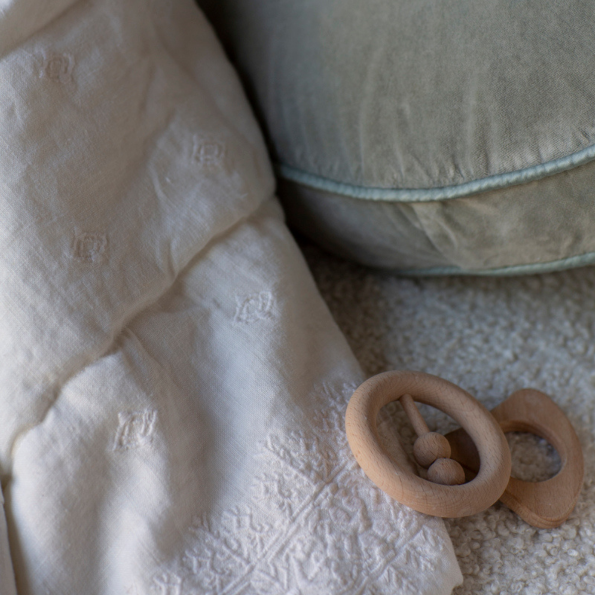: Close up of rumpled blanket in winter white highlights embroidery, shown with a pale green pillow and wooden teether toys.