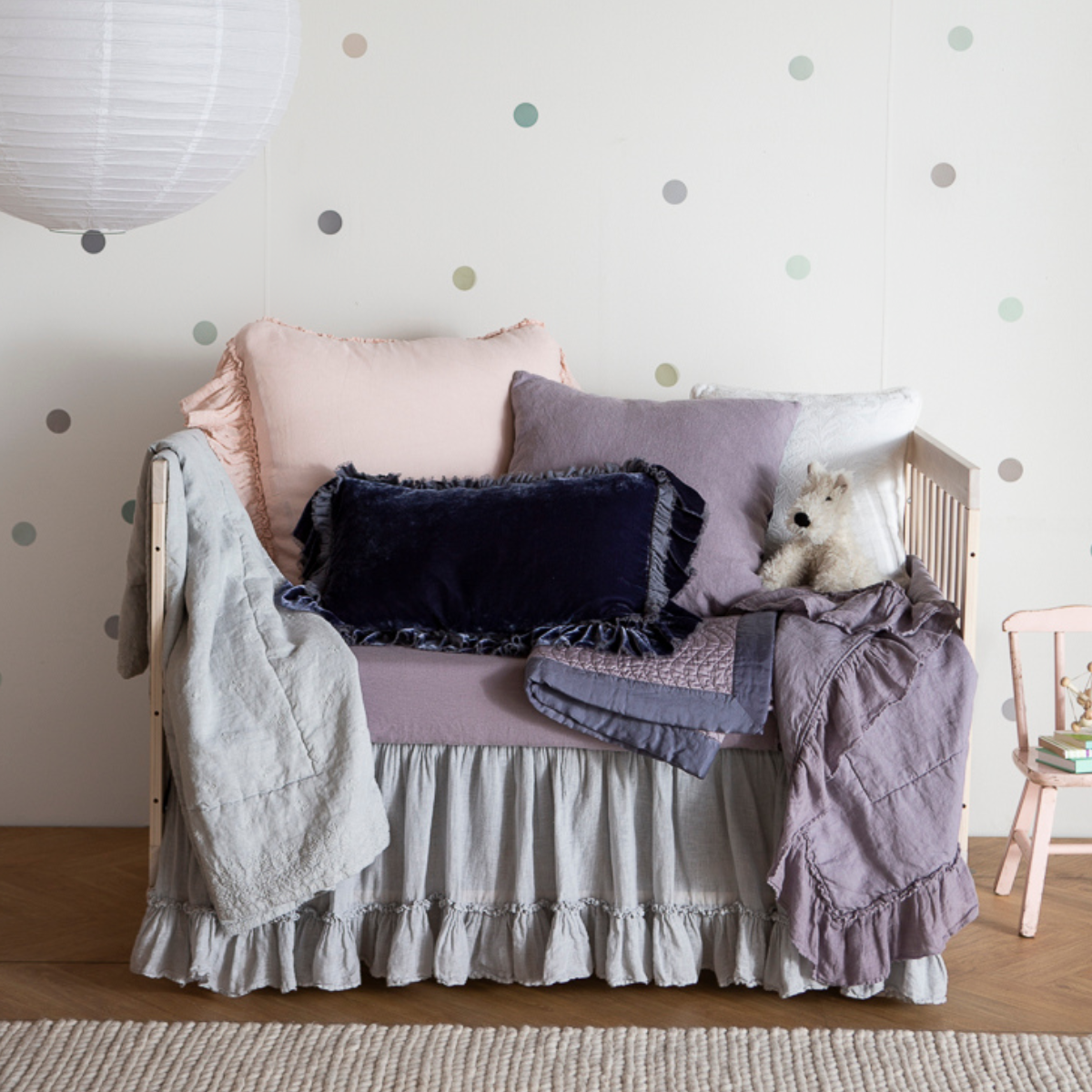 : a feminine crib dressed in french lavender, pearl and cloud with several throw pillows and a few baby blankets.
