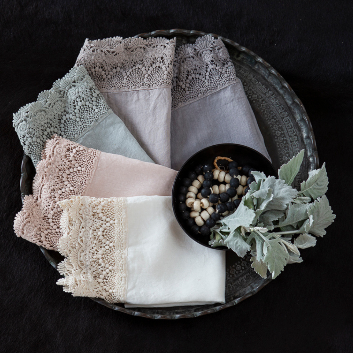 : a large tray with folded linen with lace trim in several colors with a decorative bowl of beaded jewelry and greenery.