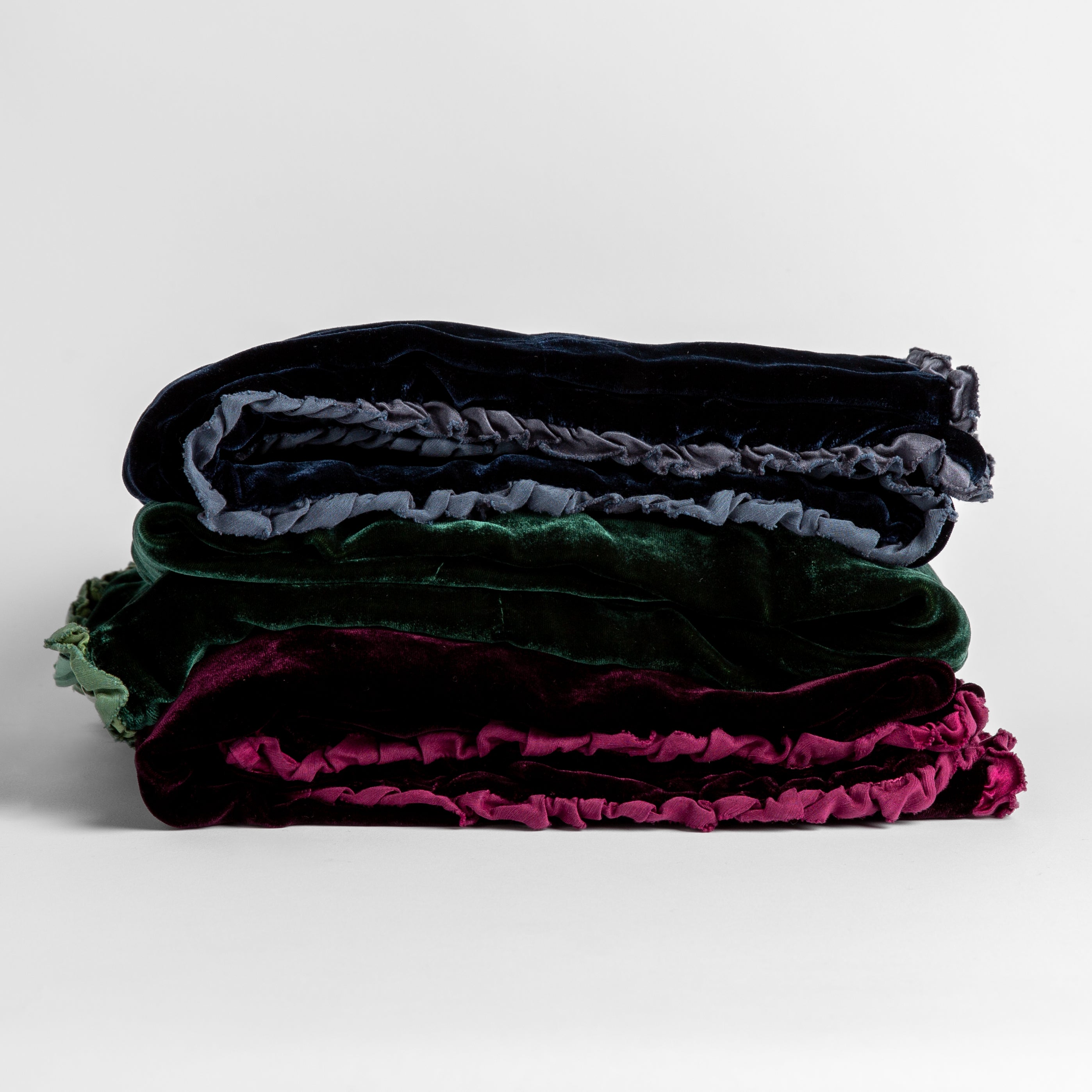 Stack of four silk velvet baby blankets with charmeuse trim shown against a white background. Blankets shown in Midnight, Jade and Fig.