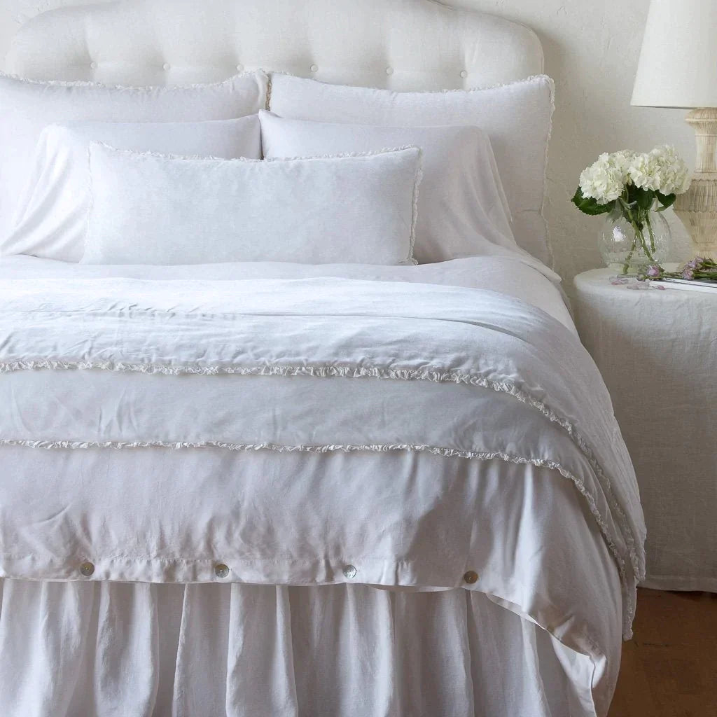 White: Silk velvet shams with a petite ruffle on a monochromatic bed, stacked behind sleeping and throw pillows - view from end of bed.