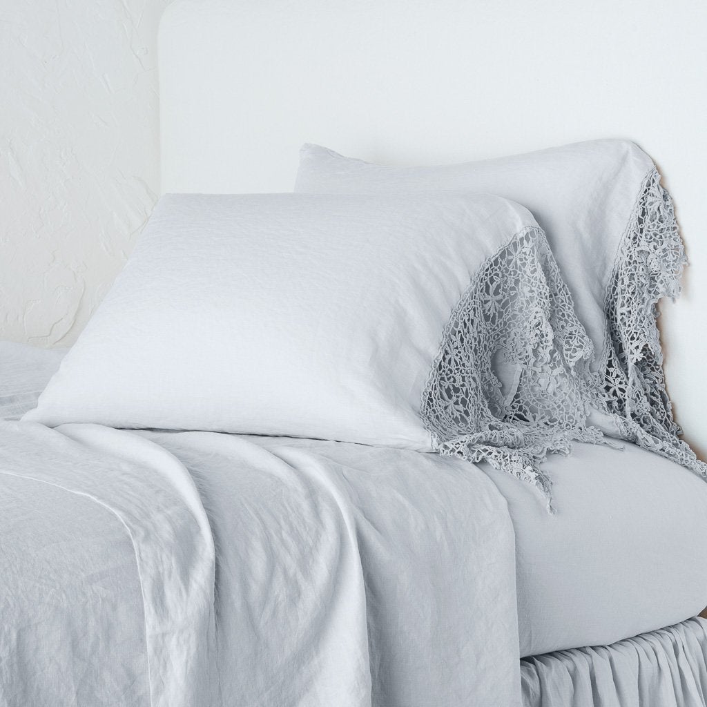 Cloud: Lace trimmed linen pillowcases leaning against a plain wall on a monochromatic bed - cropped three-quarter angle.