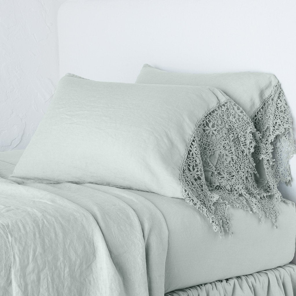 Eucalyptus: Lace trimmed linen pillowcases leaning against a plain wall on a monochromatic bed - cropped three-quarter angle.