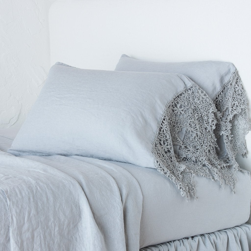 Mineral: Lace trimmed linen pillowcases leaning against a plain wall on a monochromatic bed - cropped three-quarter angle.