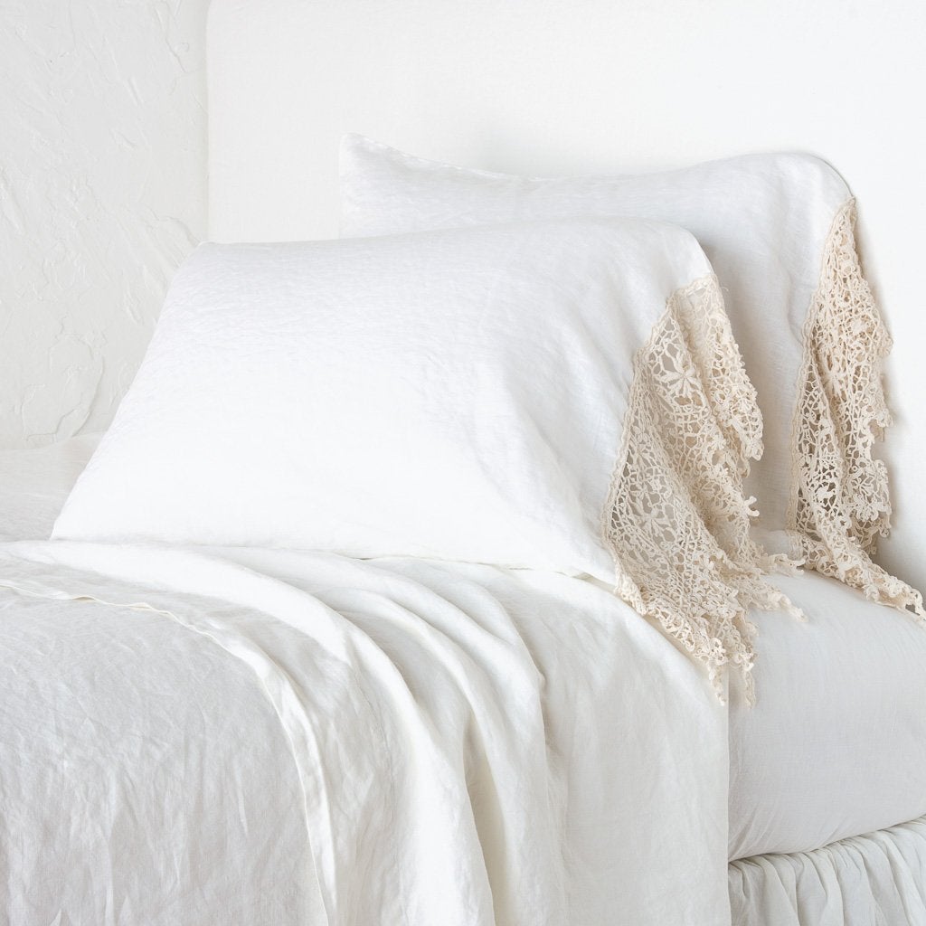 Winter White: Lace trimmed linen pillowcases leaning against a plain wall on a monochromatic bed - cropped three-quarter angle.