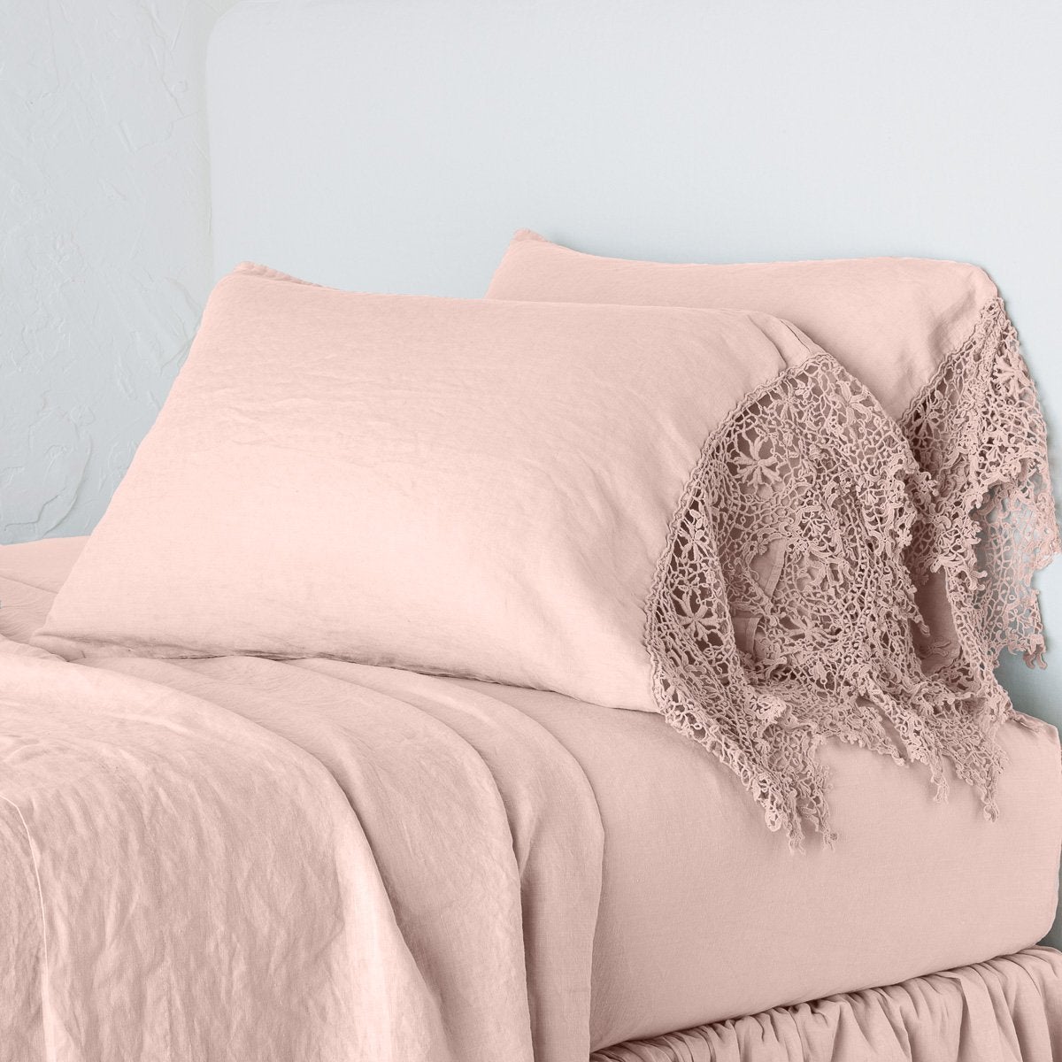 Rouge: Lace trimmed linen pillowcases leaning against a plain wall on a monochromatic bed - cropped three-quarter angle.