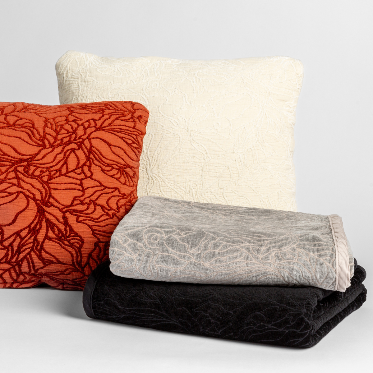 A cluster of two Georgia throw pillows displayed on their cotton jacquard side and two Georgia blankets displayed on their chenille jacquard sides, all against a white background, in the following colorways: one throw pillow in Autumn, a rich terracotta tone, another throw pillow in Winter White, a wooly and warm tone, one blanket in Fog, a neutral mid-grey, and another blanket in Corvino, a raven black tone.