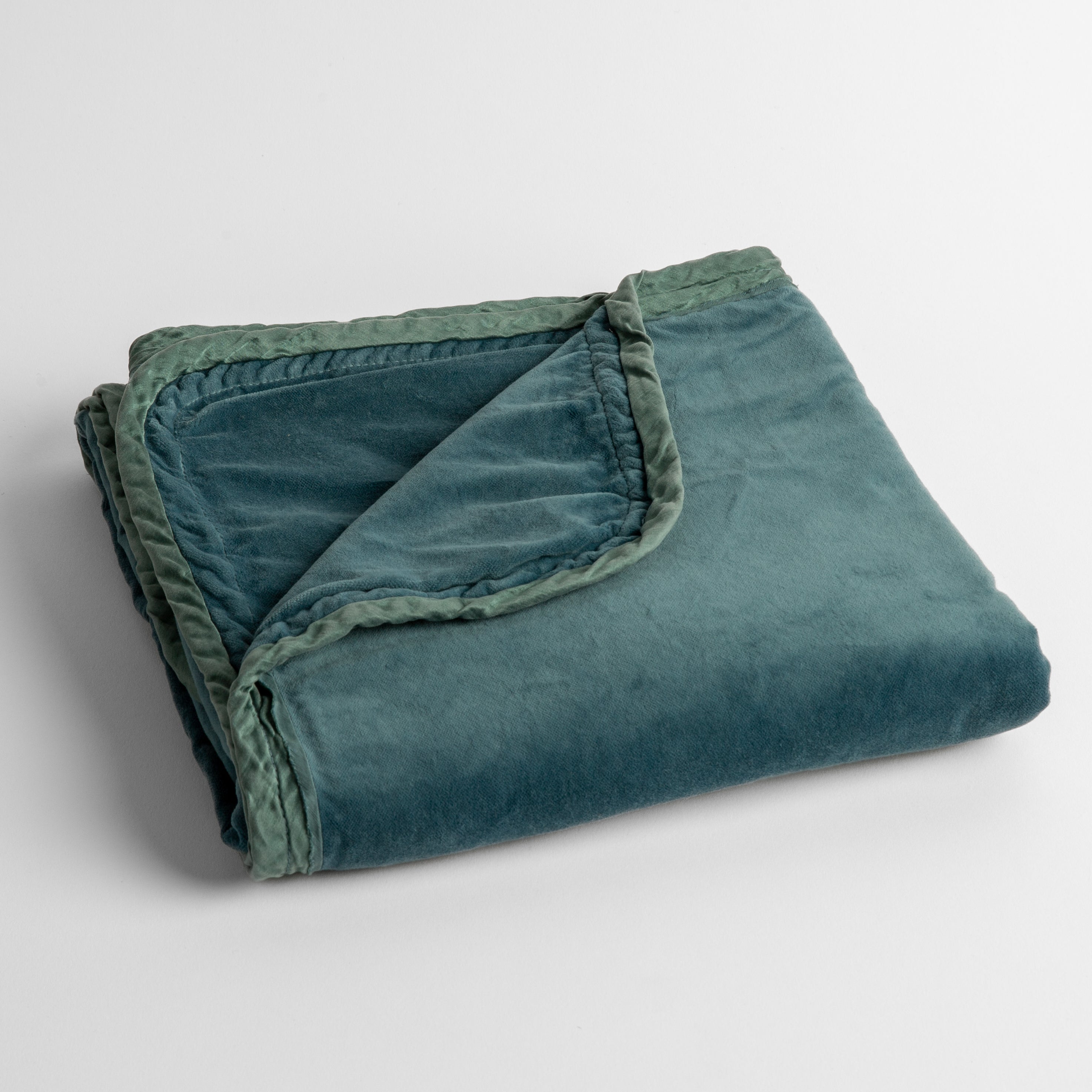 Cenote: cotton velvet baby blanket trimmed with charmeuse, folded with a covern folded back to show trim contrast; shot overhead and at a slight angle against a white background.