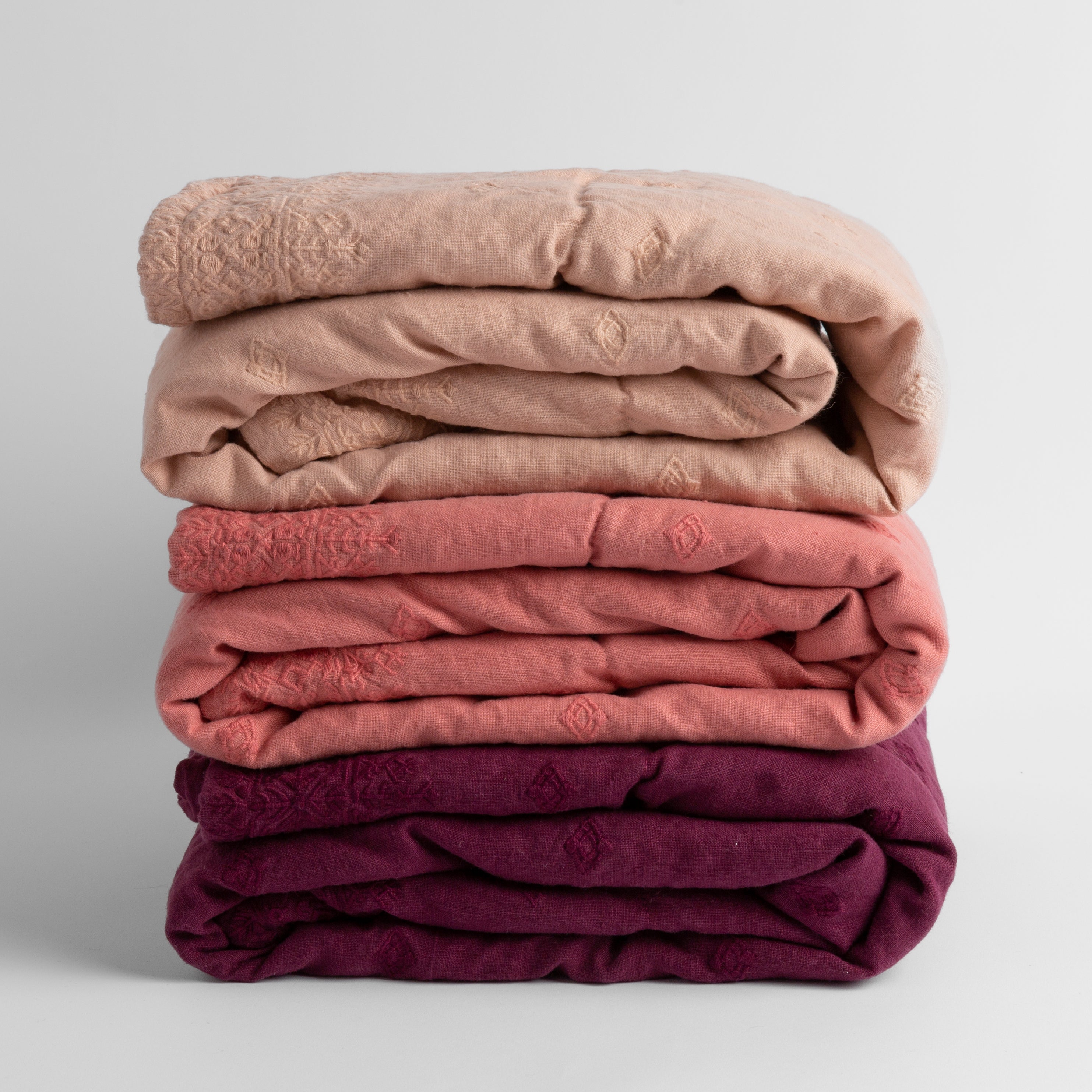 : a stack of three embroidered linen baby blankets in warm tones: pearl, poppy and fig.