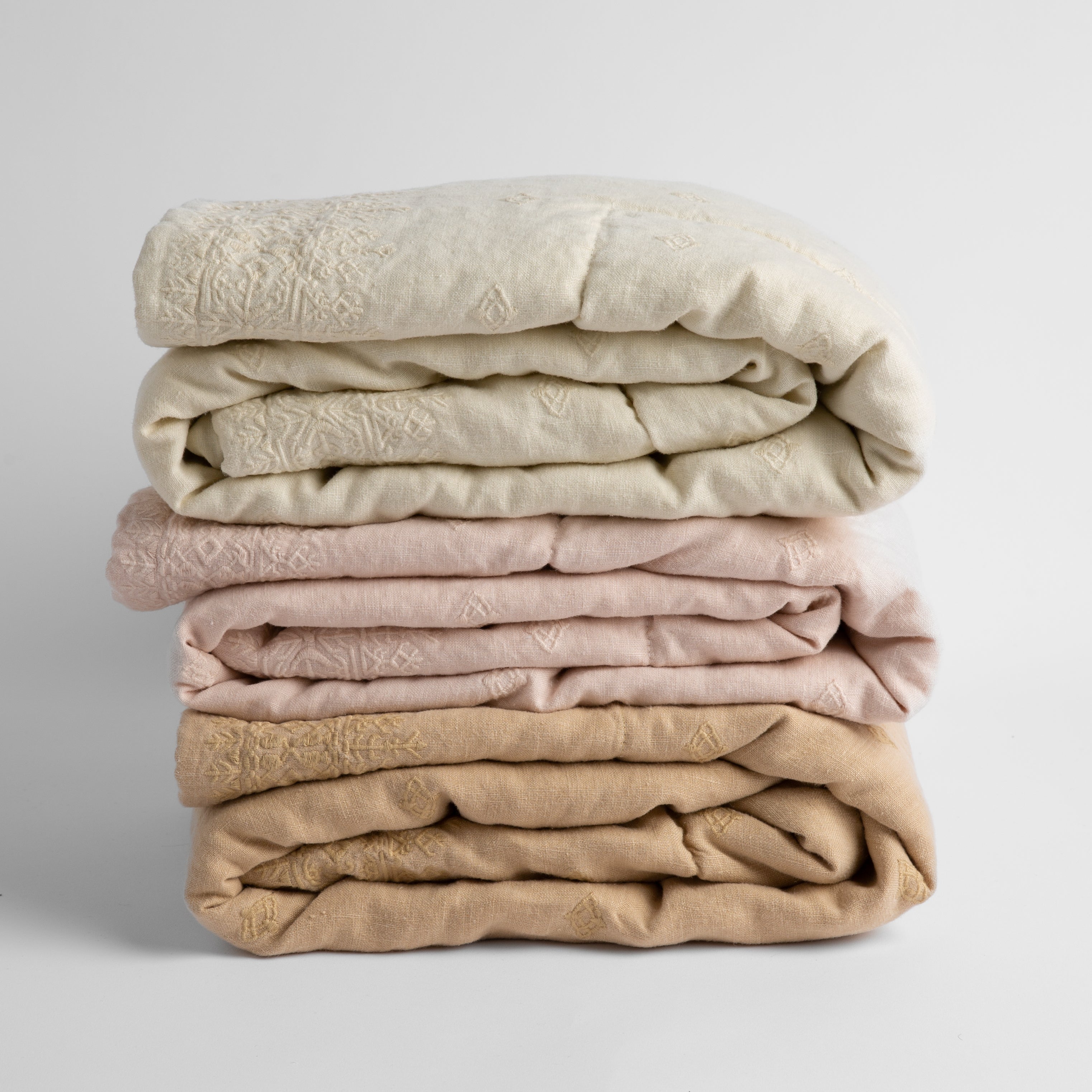 A stack of three embroidered linen baby blankets in neutral tones: parchment, pearl and honeycomb