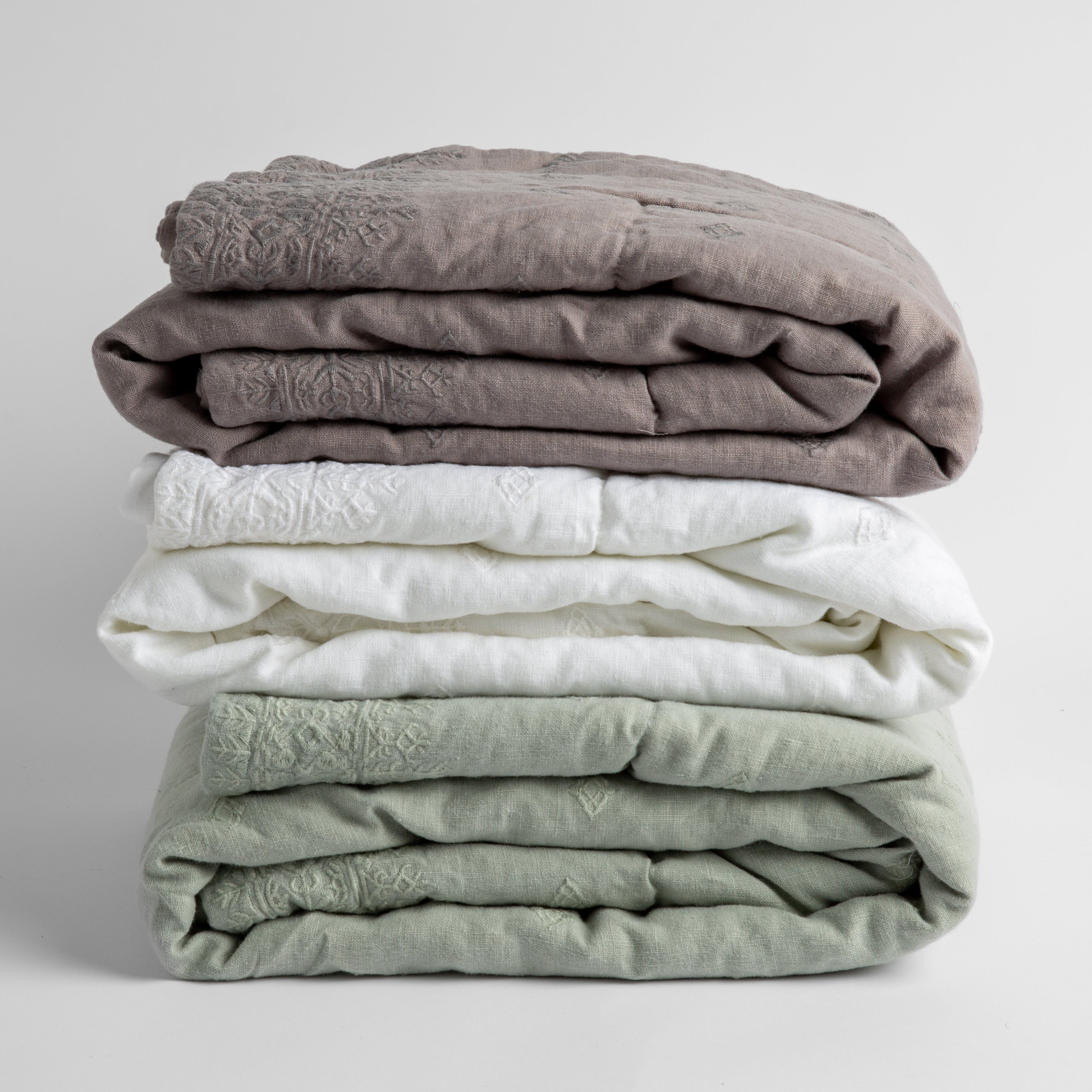 : A stack of three embroidered linen baby blankets in moonlidhgt, white and eucalyptus.