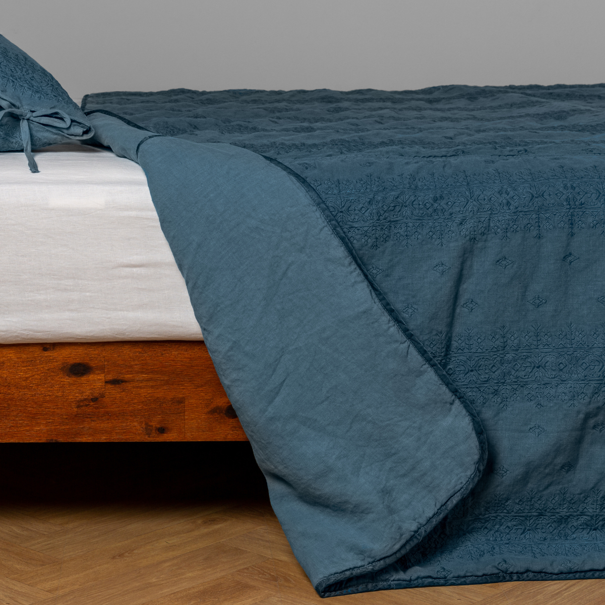 Cenote: White: embroidered bedspread with corner pulled back showing the plain linen back. Shown with a white fitted sheet and matching sham, the bedframe is slightly visible and the bed is against a white wall and on a medium wood floor.