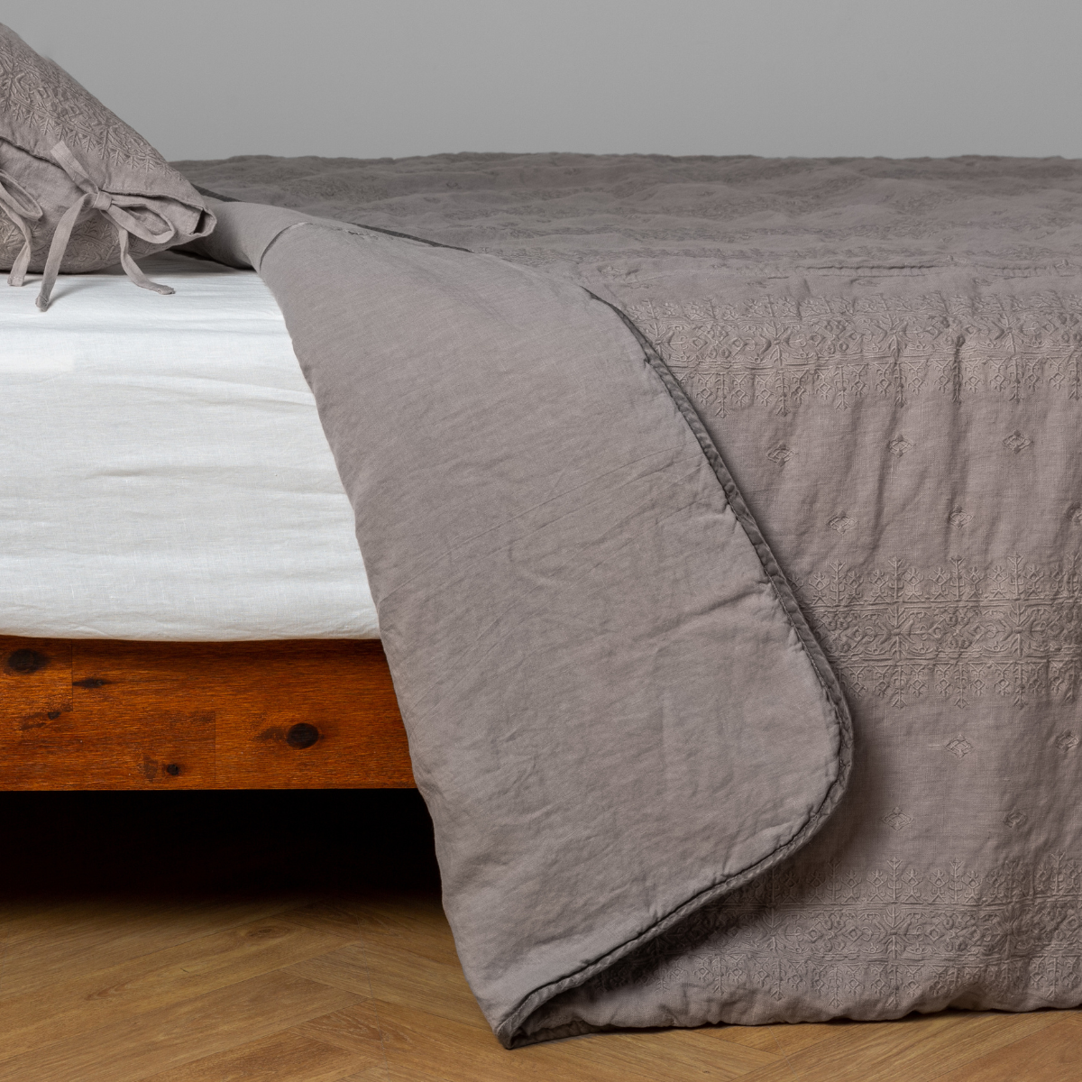 Fog: White: embroidered bedspread with corner pulled back showing the plain linen back. Shown with a white fitted sheet and matching sham, the bedframe is slightly visible and the bed is against a white wall and on a medium wood floor.