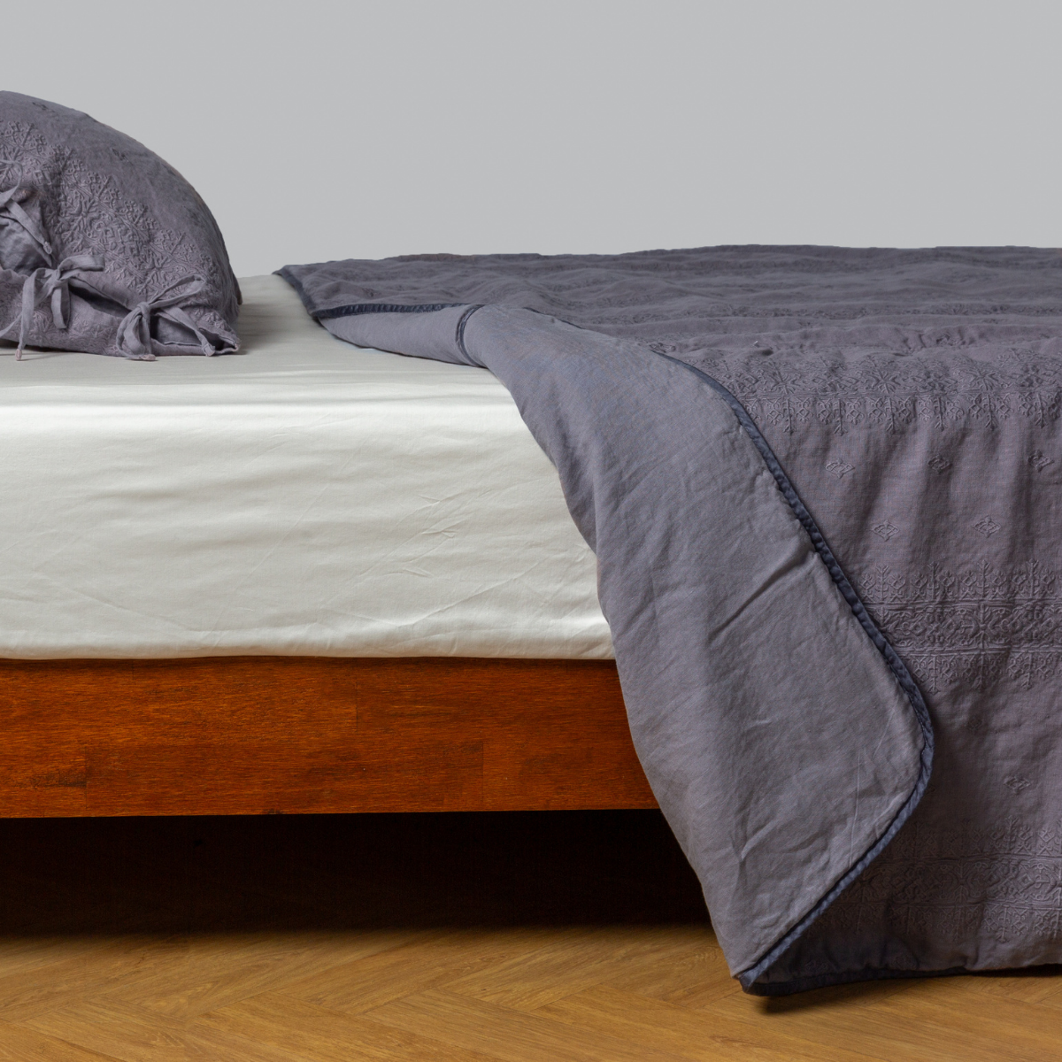 French Lavender: embroidered midweight linen bedspread, corner folded back, with matching sham on a bed with a white fitted sheet. shot from the side view, the bedframe is slightly visible against a white background and medium wood floor.