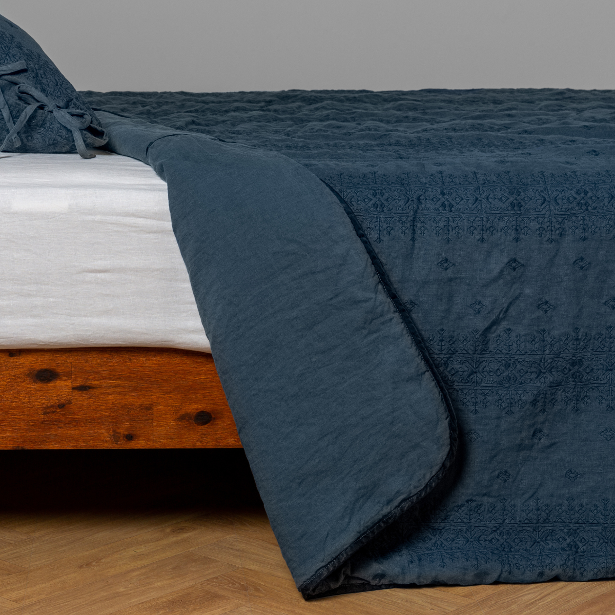 Midnight: White: embroidered bedspread with corner pulled back showing the plain linen back. Shown with a white fitted sheet and matching sham, the bedframe is slightly visible and the bed is against a white wall and on a medium wood floor.