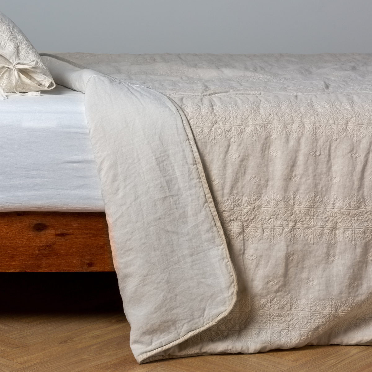 Parchment: White: embroidered bedspread with corner pulled back showing the plain linen back. Shown with a white fitted sheet and matching sham, the bedframe is slightly visible and the bed is against a white wall and on a medium wood floor.