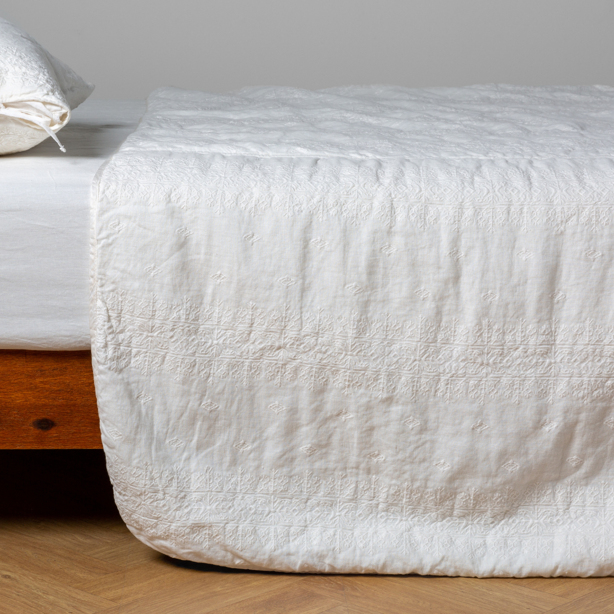 Winter White: embroidered midweight linen bedspread with matching sham on a bed with a white fitted sheet. Shot from the side view, the bedframe is slightly visible against a white background and medium wood floor.