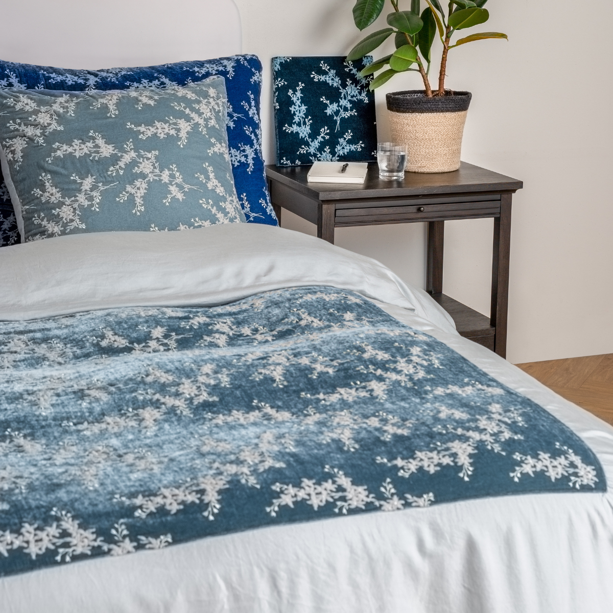 Cloud: shown from the foot of the bed, an embroidered silk velvet panel inset into a tencel duvet cover and matching shams are shown with a side table holding a notepad and pen, glass of water and potted plant.