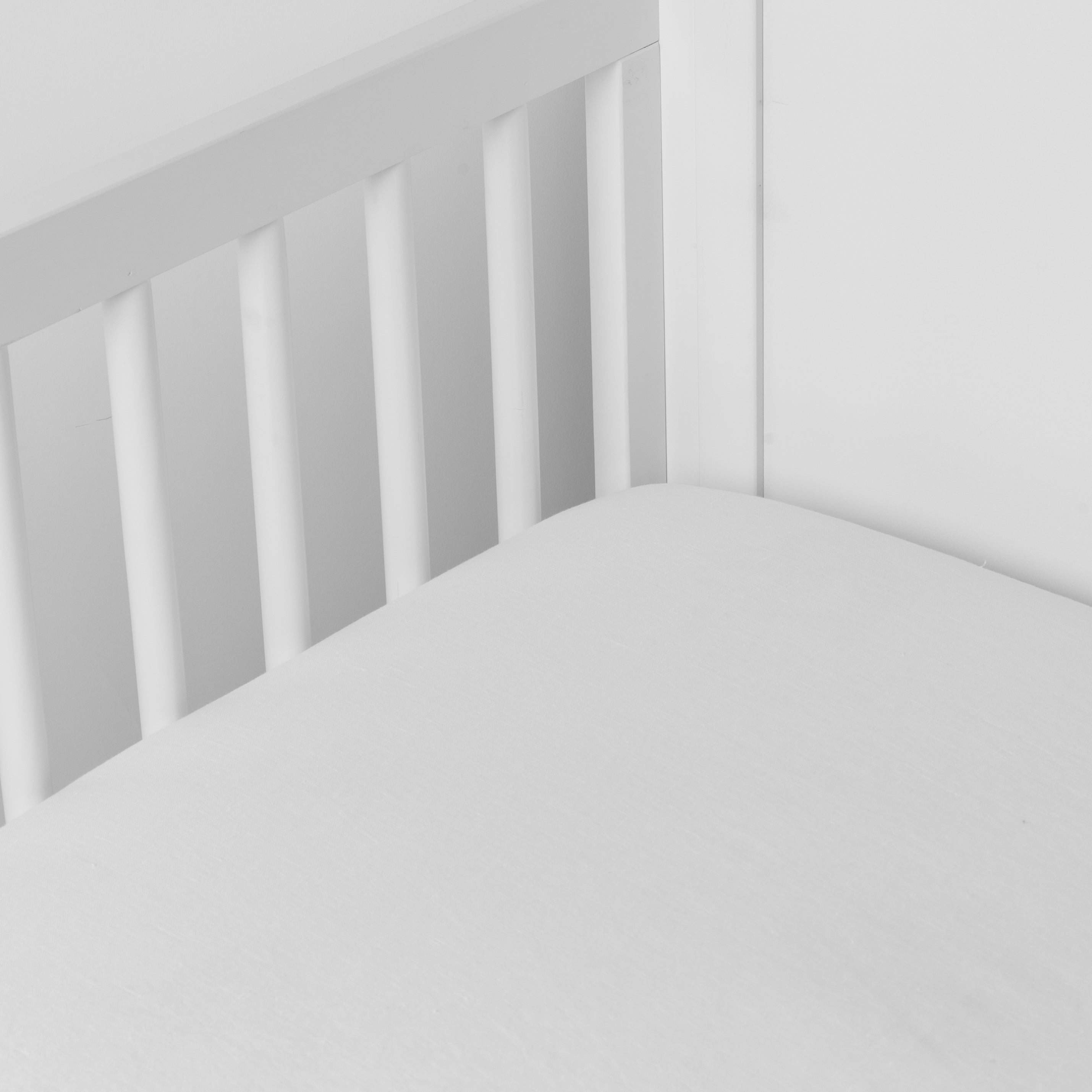 White: linen crib sheet on the mattress shot slightly overhead into the corner of a white crib. against a white background.