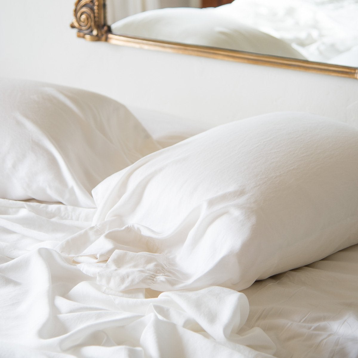 : Madera Luxe sleeping pillows and sheeting in winter white, rumpled against a white wall with an antique gold frame mirror partially visible.