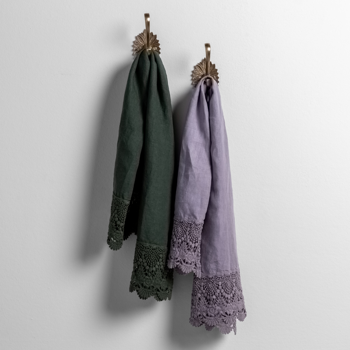 : mattine lace trimmed linen guest towels in juniper and french lavender hanging from decorative towel hooks against a white wall.