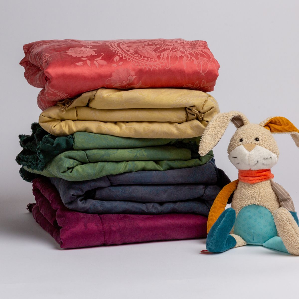 : a stack of bold rainbow colored silk and tencel™ baby blankets shown with a colorful stuffed bunny.