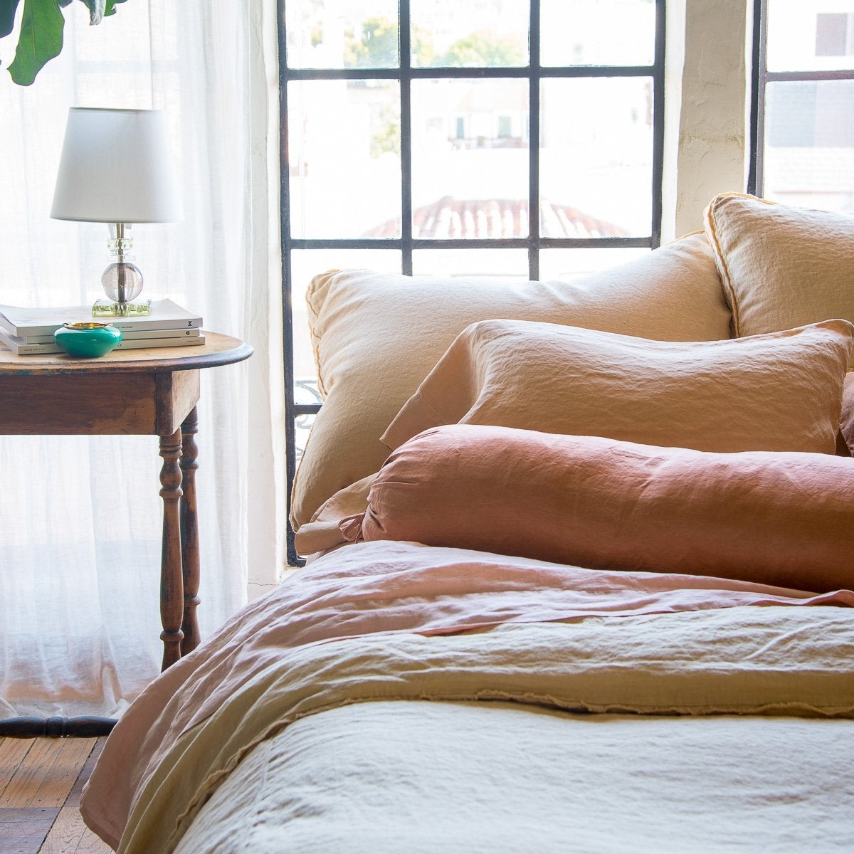 : Paloma sleeping pillow and bolster on linen bedding, in soft pink and gold tones against big windows - cropped end of bed view.