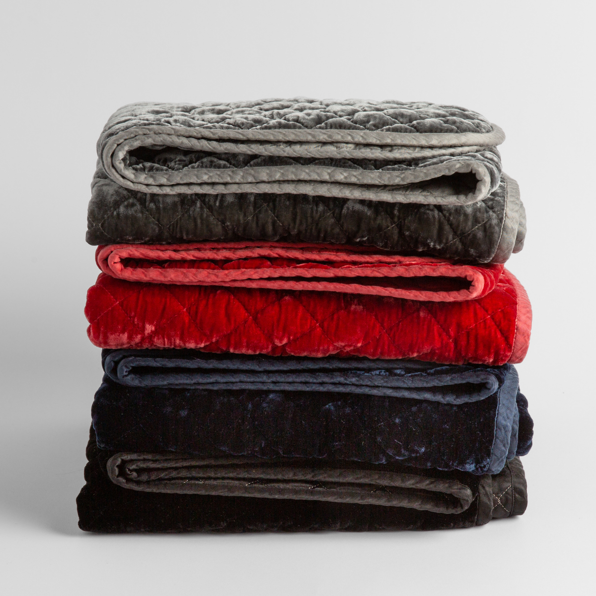 stack of 4 quilted silk velvet baby blankets in fog, poppy, midnight and moonlight against a white background.