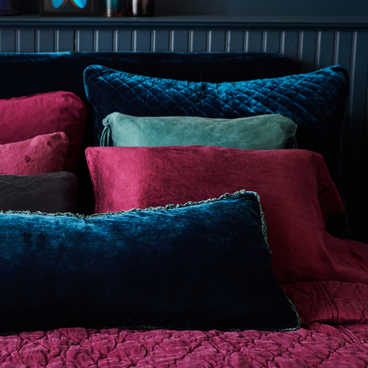 : Silk velvet sham in cenote, with matching lumbar pillow, layered with fig and cenote silk charmeuse against a moody dark headboard and wall - cropped end of bed view.
