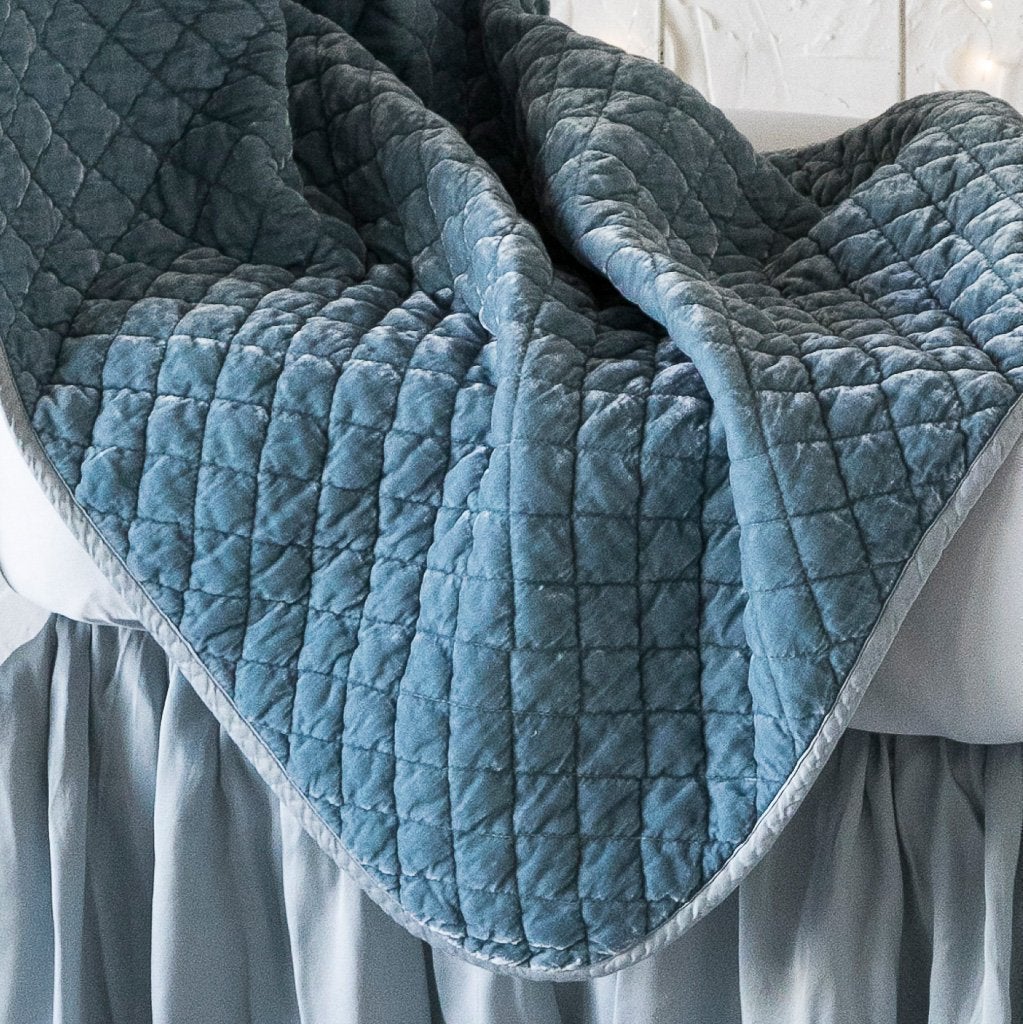 : Close up of silk velvet quilted baby blanket in cloud, draped over a monochromatic crib, showcasing the satin edge trim.