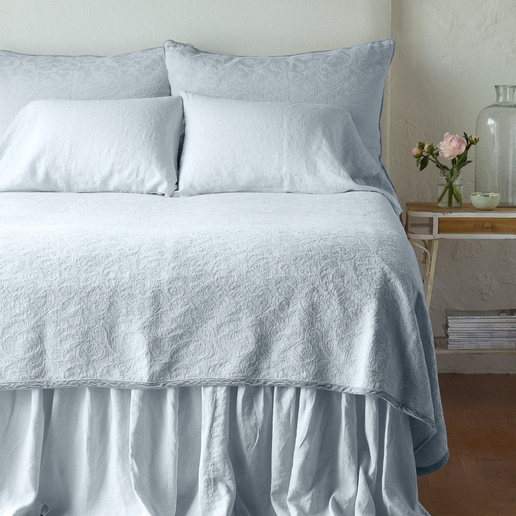 Cloud: The Adele coverlet in cloud, viewed from the foot of the bed, with white bed skirt and pillows visible. The headboard is plain light color, and part of a bedside table is visible with flowers, a vase and some books. against a plain white wall.
