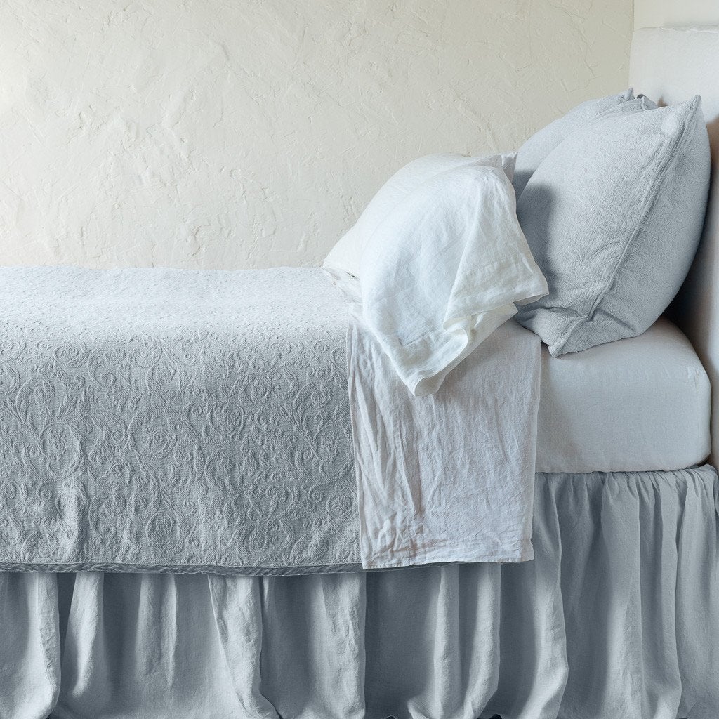 Cloud: The Adele coverlet in cloud, on a bed viewed from the side against a plain white wall. The bed is styled neatly with the coverlet folded back to reveal white sheets, bed skirt and pillows.