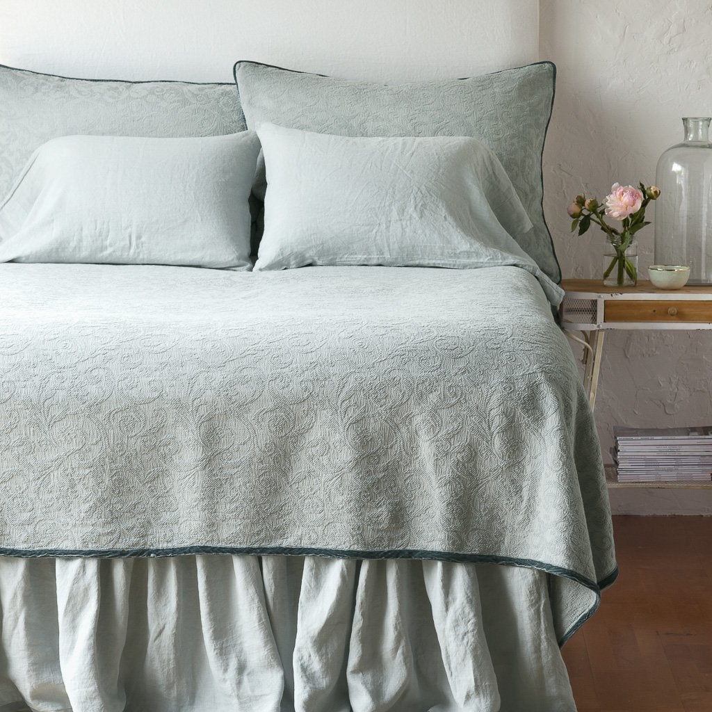 Eucalyptus: The Adele coverlet in eucalyptus, viewed from the foot of the bed, with white bed skirt and pillows visible. The headboard is plain light color, and part of a bedside table is visible with flowers, a vase and some books. against a plain white wall.