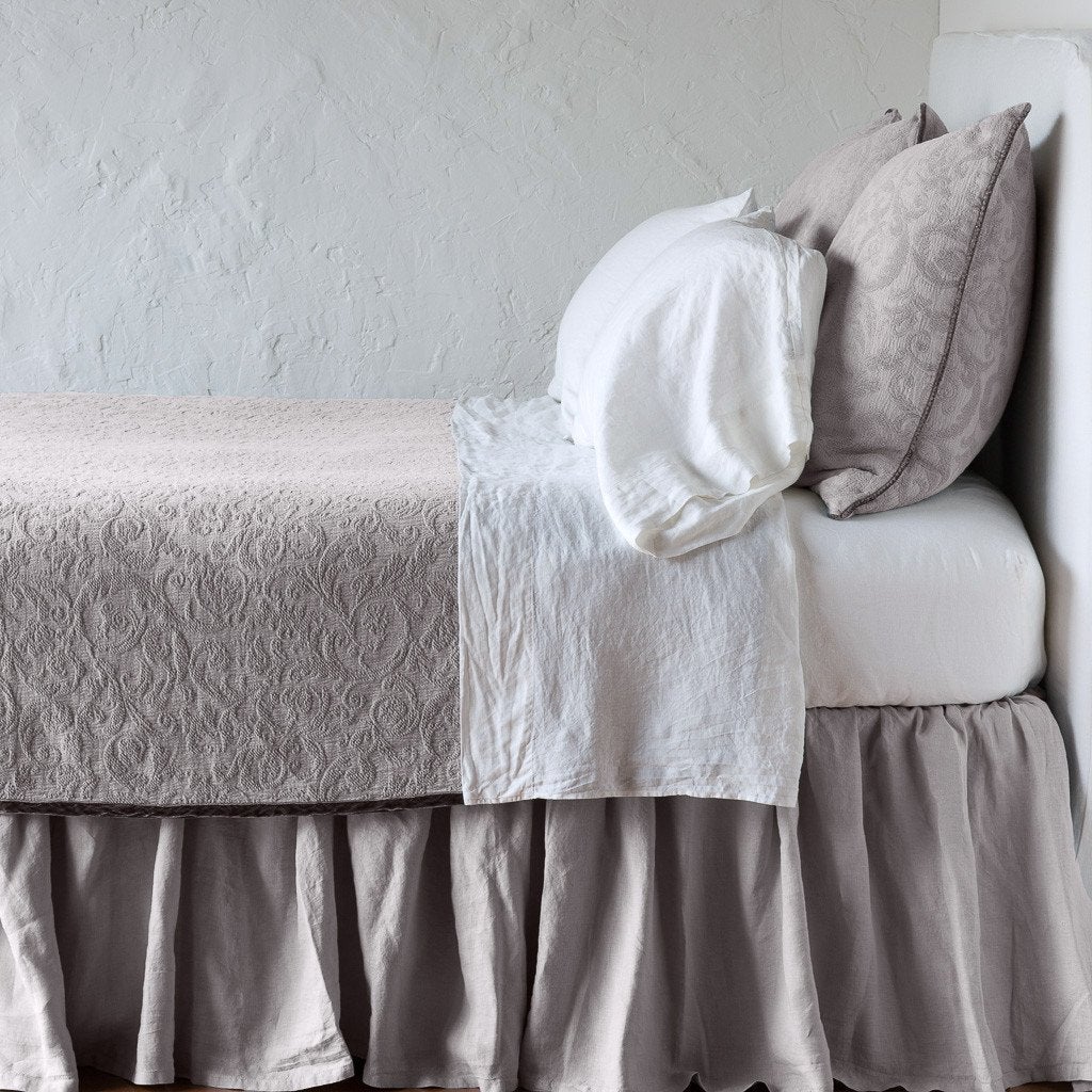Fog: The Adele coverlet in fog, on a bed viewed from the side against a plain white wall. The bed is styled neatly with the coverlet folded back to reveal white sheets, bed skirt and pillows.