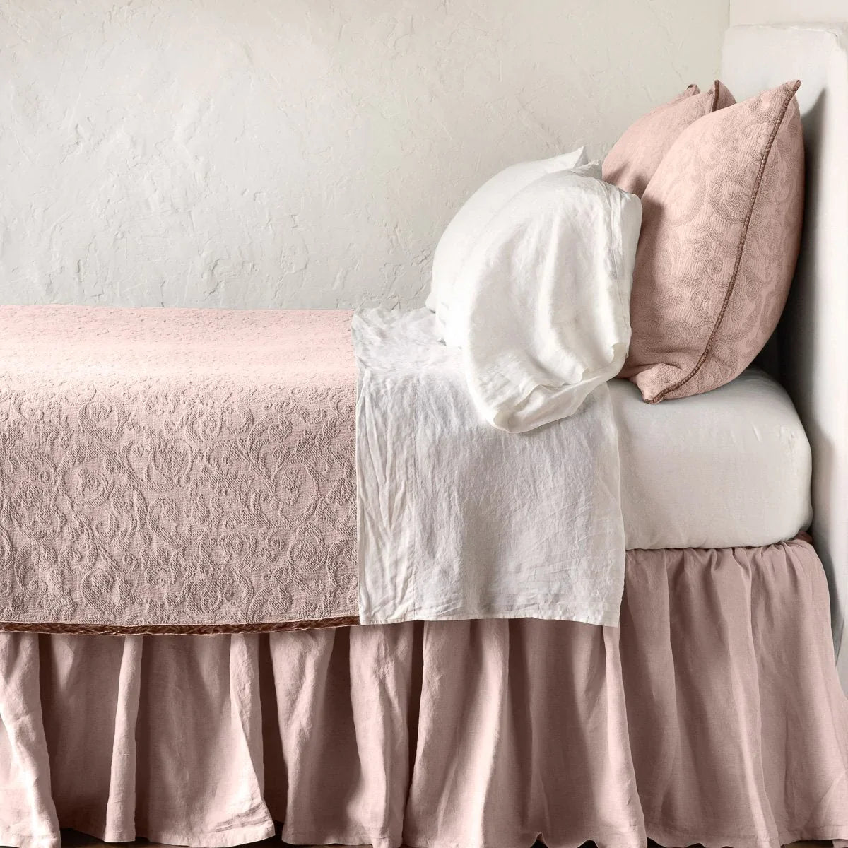 Rouge: The Adele coverlet in rouge, on a bed viewed from the side against a plain white wall. The bed is styled neatly with the coverlet folded back to reveal white sheets, bed skirt and pillows.