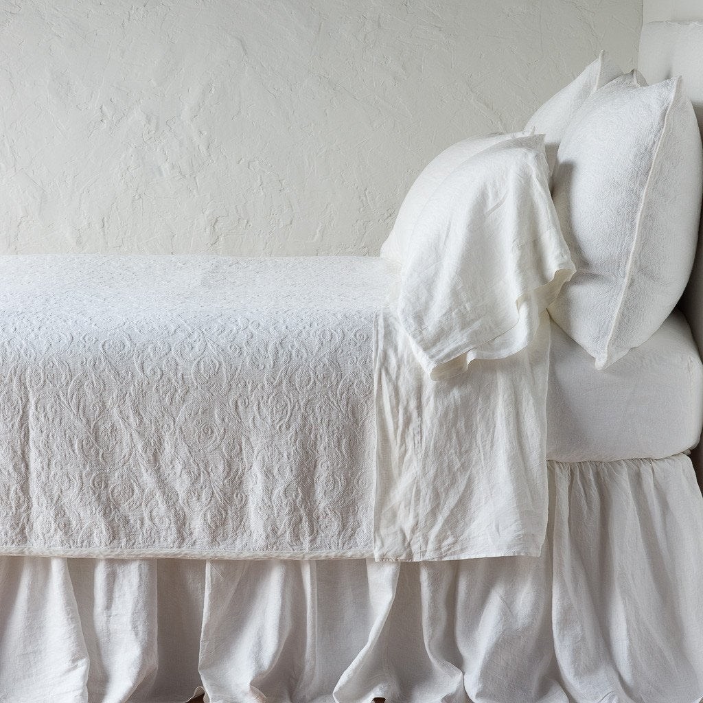 Winter White: The Adele coverlet in winter white, on a bed viewed from the side against a plain white wall. The bed is styled neatly with the coverlet folded back to reveal winter white sheets, bed skirt and pillows.