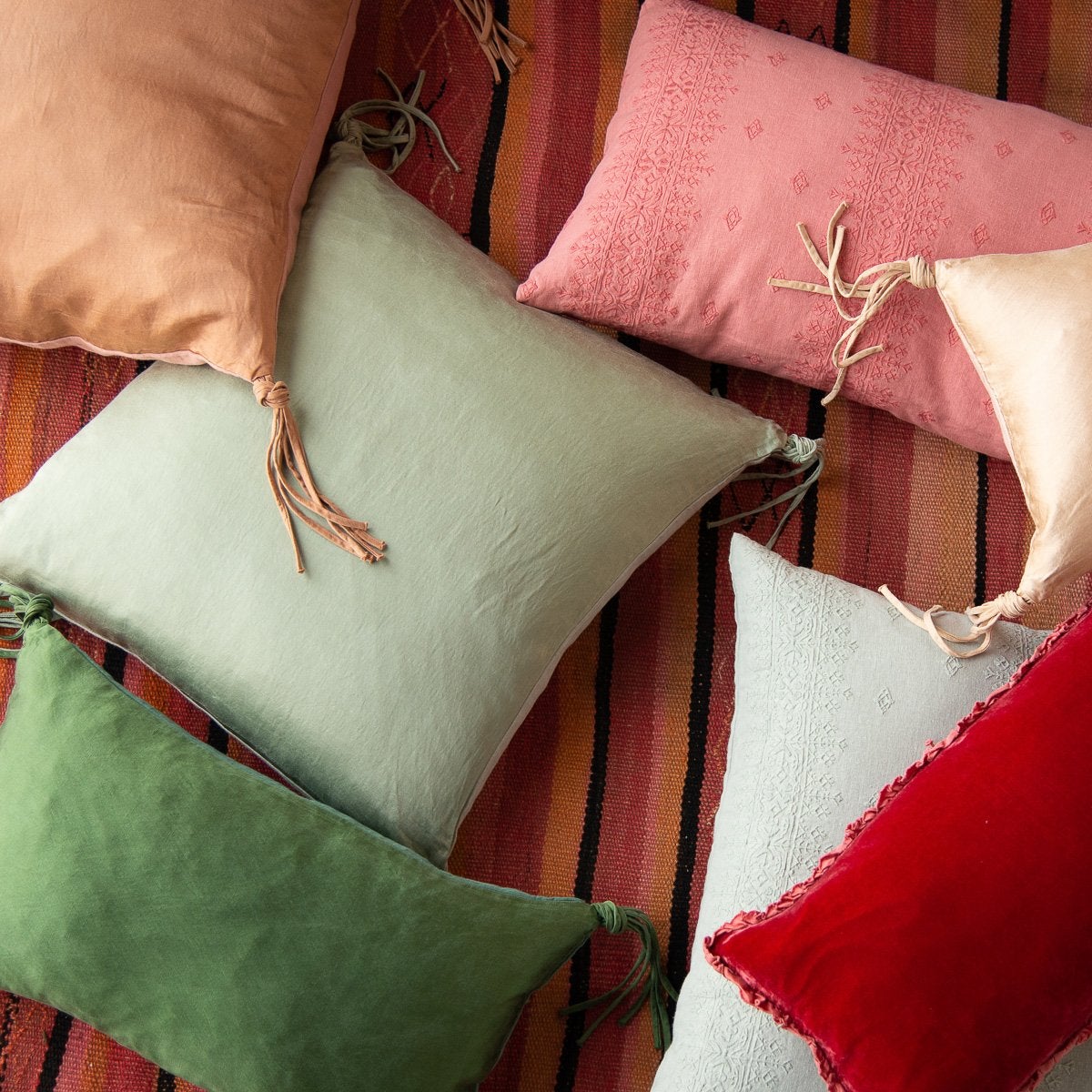 : Ines, silk charmeuse, and silk velvet throw pillows in pink and green tones, scattered on a boldly striped rug - overhead view.