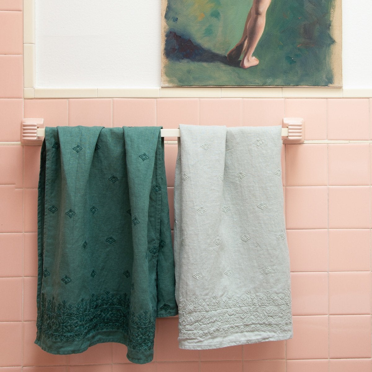 : Ines guest towels in jade and eucalyptus, rumpled on a towel bar against a pink tiled wall, under on-tone art.