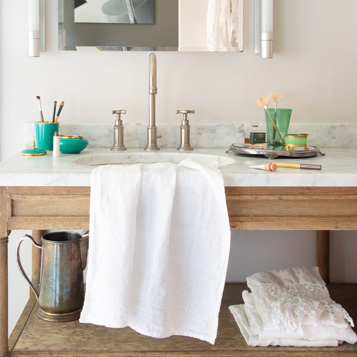 : White Ines guest towel draped over a marble and wood bathroom vanity, with makeup accessories in green glass jars.