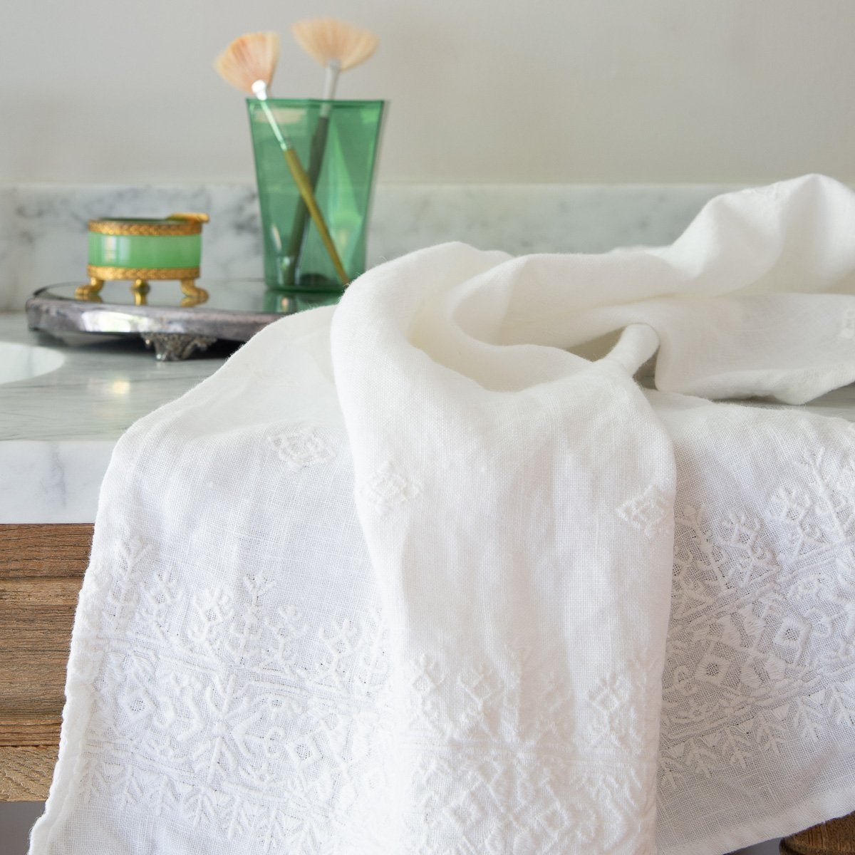 : White Ines guest towel draped over a marble bathroom counter with green glass accessories in the background.