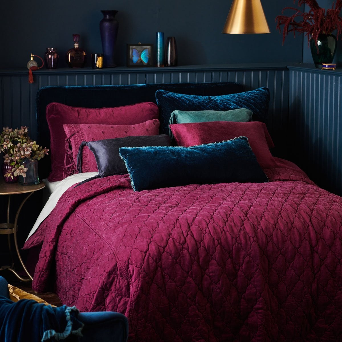 : Carmen lumbar pillow in cenote, on a bed with deep fig and dark blue tones. Walls and art/accessories are a moody dark blue.