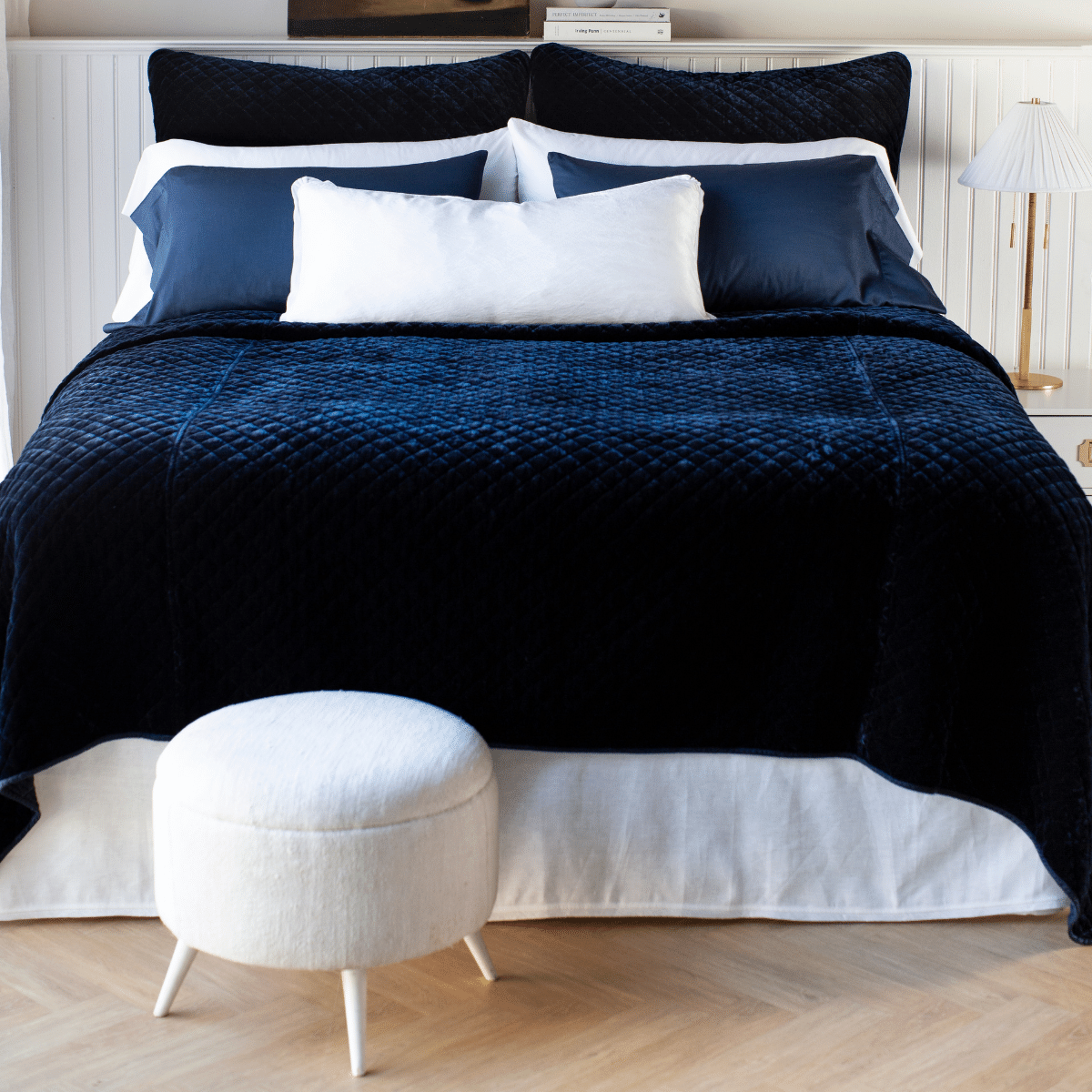 : Silk velvet quilted shams and matching coverlet in deep midnight blue, layered simply with white and blue sleeping pillows and a white lumbar pillow - end of bed view.